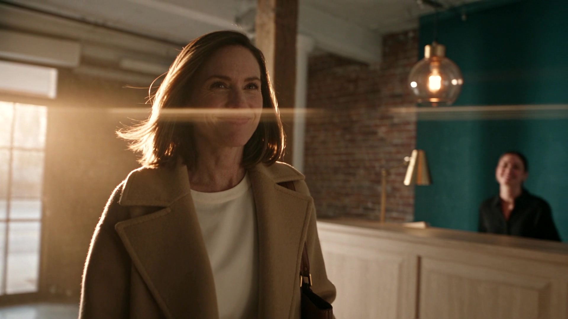 Film of models: a smiling woman in a camel coat arrives at a sunlit reception desk