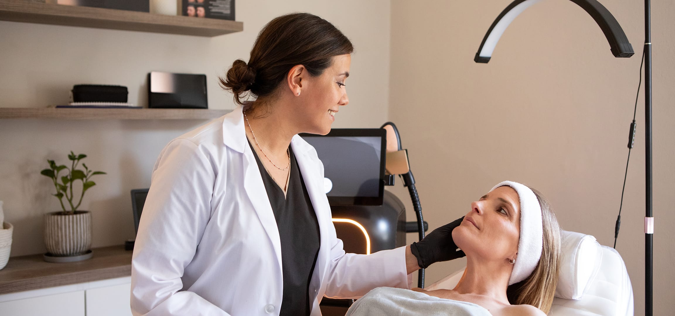 Photograph of models: a clinician in a white coat prepares a reclining patient for a skin treatment in a bright treatment room