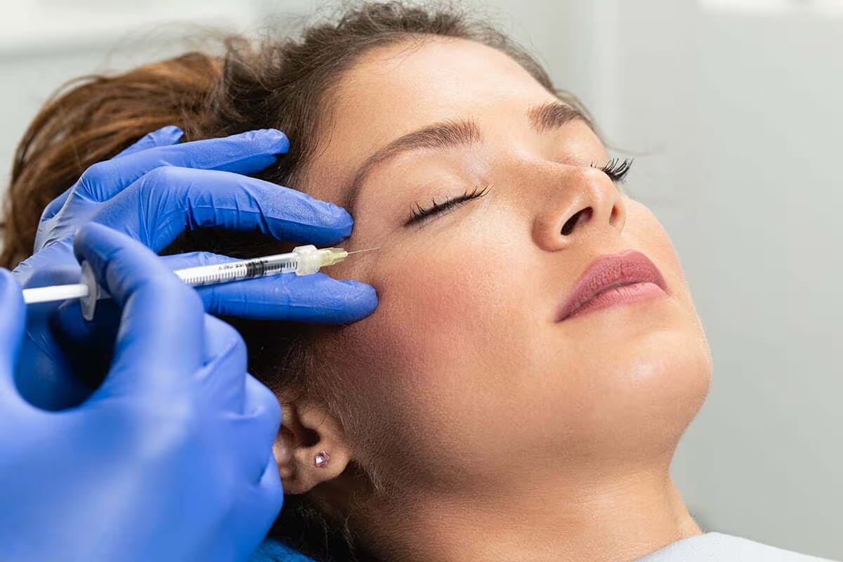 Photograph of a model: a gloved injector places a fine needle at the outer corner of a relaxed patient's eye
