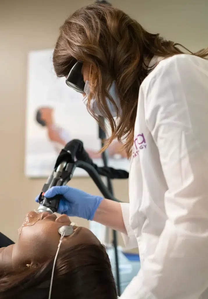 Photograph of models: a clinician in protective glasses treats a reclining patient with a laser hair removal handpiece
