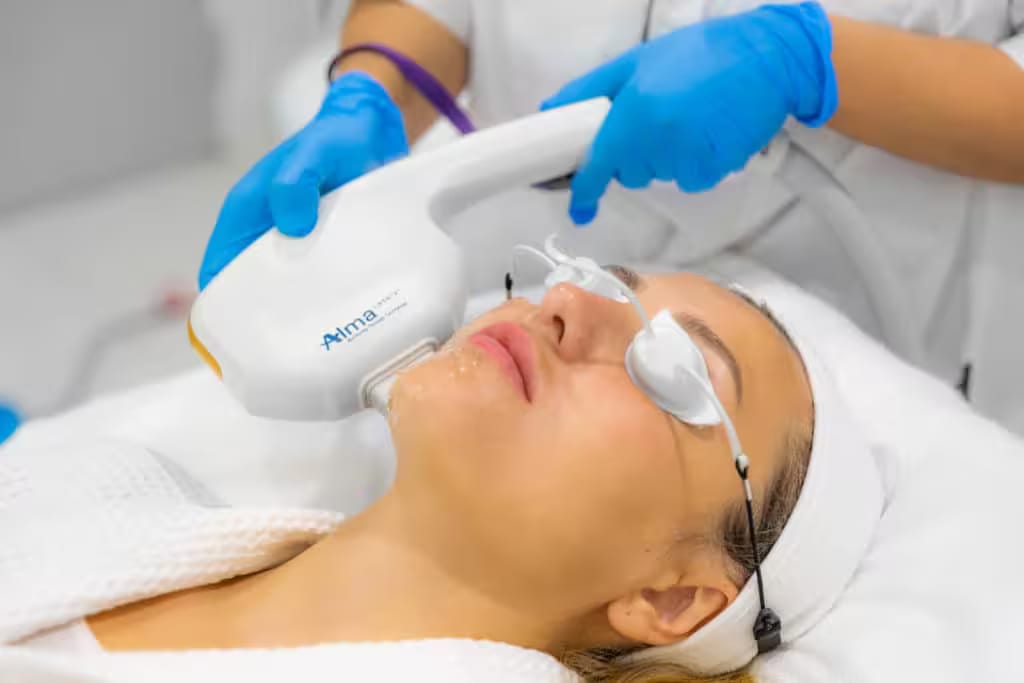 Photograph of models: a gloved clinician moves a laser handpiece across the cheek of a patient wearing eye shields